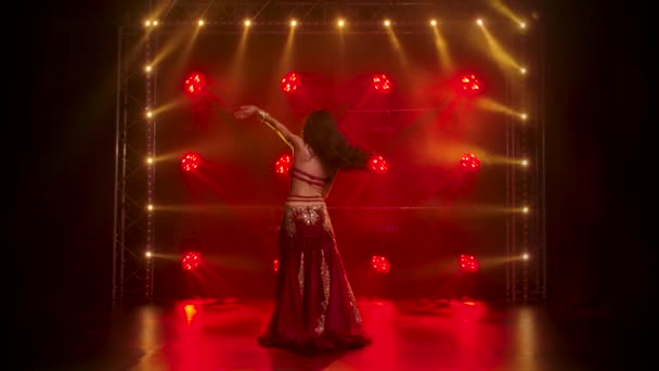 Kobieta w egzotycznym stroju wykonuje taniec brzucha porusza pół nagie ciało. Kręcony w ciemnym studiu z dymem i czerwonym neonem. Zwolniony ruch. — Wideo stockowe