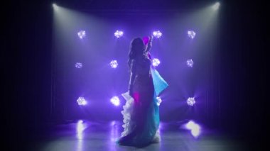 Beyaz doğulu bir kostüm giymiş, ipek kumaşla dans eden kadın dansçı. Duman ve mavi ışıklı karanlık bir stüdyoda çekildi. İnce, esnek bir vücudun siluetleri. Yavaş çekim.