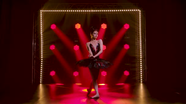 Půvabná baletka v elegantním obrazu černé labutě. Mladá krásná dívka v černé sukničce s červenými flitry a korunou. Natočeno v tmavém studiu s kouřem a červeným neonovým osvětlením. Zpomalený pohyb. — Stock video