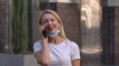 Düşük korumalı tıbbi maske takmış güzel bir genç kadının portresi akıllı telefonuyla konuşuyor. COVID-19 önleyici önlemler. Yan görüş. Ağır çekim. Kapat..