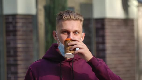 Porträtt av ung man sänker en medicinsk mask och dricker kaffe medan du njuter av en aromatisk dryck. Förebyggande åtgärder enligt COVID-19. Sakta i backarna. Närbild. — Stockvideo