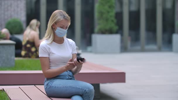 Portrett av en pen ung kvinne i medisinsk maske sittende på en benk og skrive en beskjed på sin smarttelefon eller chatte på sosiale nettverk. COVID-19: forebyggende tiltak. Langsom bevegelse. Lukk. – stockvideo