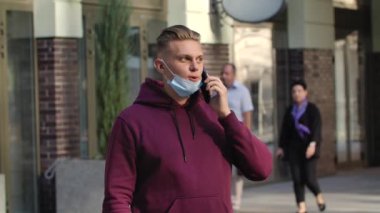 Düşük tıbbi maskeli genç adamın portresi onun akıllı telefonuyla konuşuyor. COVID-19 önleyici önlemler. Ağır çekim. Kapat..