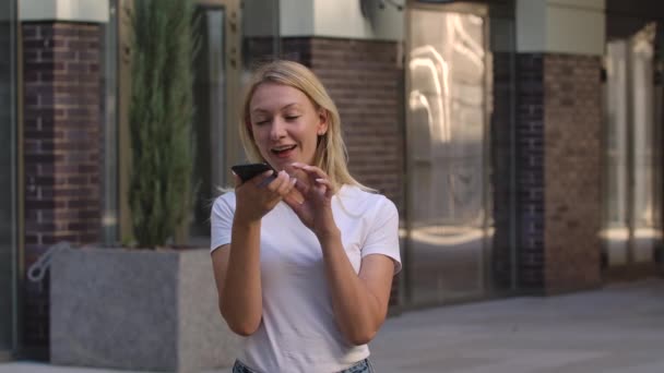 Retrato de uma mulher muito jovem falando em seu smartphone em alto-falante ou grava uma mensagem de voz. Loira em uma blusa branca ao ar livre no fundo da rua turva. Movimento lento. Fechar. — Vídeo de Stock