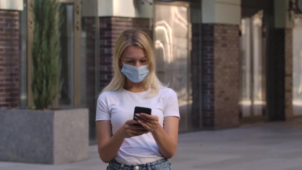 Portrait de jolie jeune femme en masque médical de protection est SMS message sur son smartphone. COVID-19 mesures préventives. Au ralenti. Gros plan. — Video