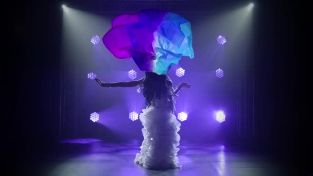 Bailarina del vientre femenina en un traje oriental blanco bailando con tela de seda. Grabado en un estudio oscuro con humo e iluminación de neón azul. Siluetas de un cuerpo delgado y flexible. Movimiento lento. — Vídeo de stock