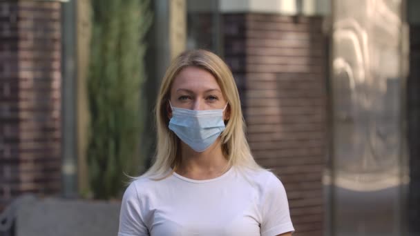Egy csinos fiatal nő arcképe orvosi maszkban, aki a kamerába néz. Szőke, fehér blúzban a szabadban, a háttérben elmosódott épületben. COVID-19 megelőző intézkedések. Lassú mozgás. Közelről.. — Stock videók