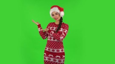Noel Baba şapkalı tatlı kız bir şeyi işaret ediyor sonra başını sallıyor ve başparmağını sallıyor. Yeşil Ekran