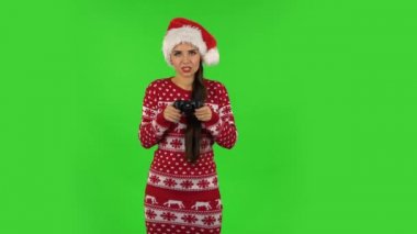 Noel Baba şapkalı tatlı kız kablosuz kumandayı kullanarak video oyunu oynuyor ve zaferle seviniyor. Yeşil Ekran