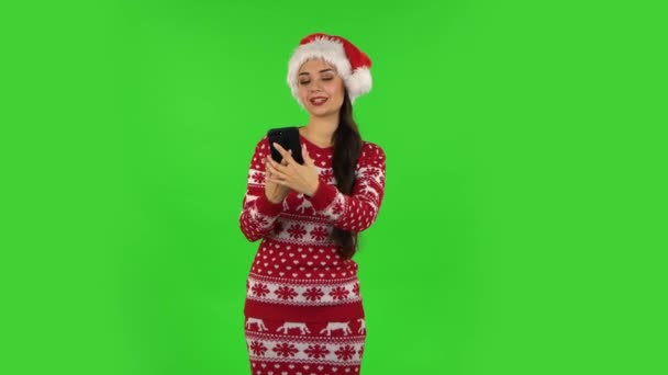 Милая девочка в шляпе Санта-Клауса делает селфи на мобильном телефоне, а затем смотрит фотографии. Зеленый экран — стоковое видео