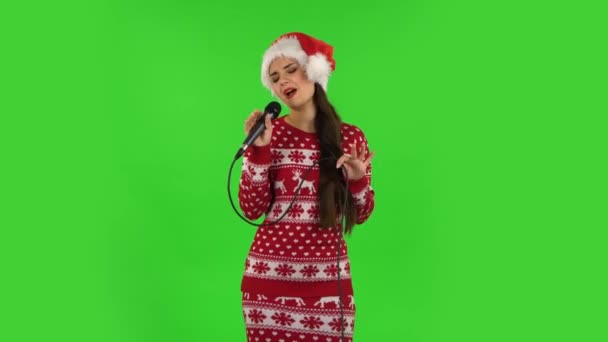 Милая девочка в шляпе Санта-Клауса поет в микрофон и движется под ритм музыки. Зеленый экран — стоковое видео
