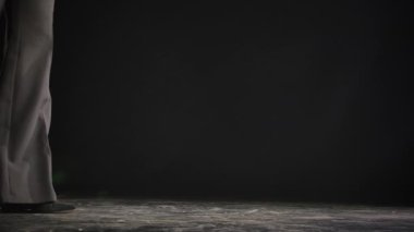 Zarif pantolonlu kadın bacakları ve stüdyoda yerde yürüyen siyah topuklu ayakkabılar. Bacaklar ve ayakkabılar siyah arkaplanda ağır çekimde kapanır..
