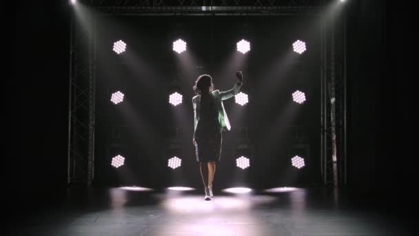 Een aantrekkelijke brunette van middelbare leeftijd loopt in een donkere studio tegen de achtergrond van neonverlichting. Een vrouw in een jurk en witte schoenen loopt en kijkt intens naar beneden. Silhouet. Langzame beweging. — Stockvideo