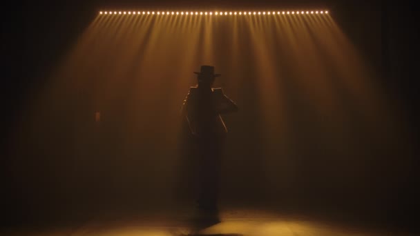 Silhouet van een aantrekkelijke vrouw in een broekpak en hoed genietend van een flamenco dans in het donker tussen de studioflitsers. Zwarte achtergrond met geel hoogtepunt. Langzame beweging. — Stockvideo