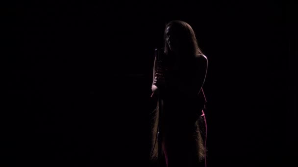 Η σιλουέτα μιας νεαρής τραγουδίστριας με μακριά μαλλιά τραγουδά σε ένα vintage μικρόφωνο στο σκοτάδι. Κοντινό πλάνο σε σκοτεινό στούντιο με οπίσθιο φωτισμό. — Αρχείο Βίντεο