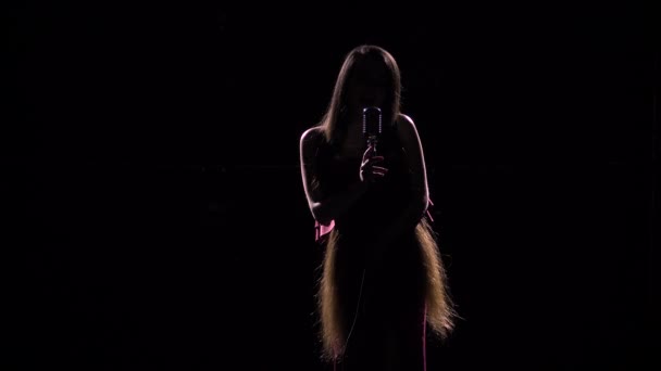 Beautiful woman with long hair sings into a vintage microphone in the dark. Female silhouette close up. — Stock Video