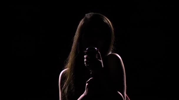 Una silueta oscura de una joven atractiva que canta en un micrófono vintage. Grabado en un estudio oscuro con luz de fondo. Hombros y cara de cerca. — Vídeos de Stock