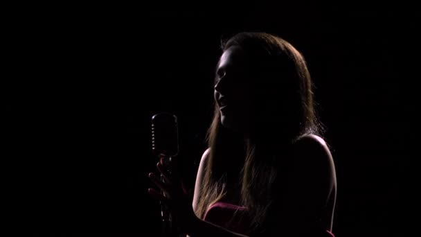 Πλευρική άποψη μιας νεαρής ελκυστικής γυναίκας που τραγουδάει σεξουαλικά σε ένα παλιό μικρόφωνο. Πυροβολήθηκε σε σκοτεινό στούντιο με πίσω φως. Θηλυκή σιλουέτα κοντά. — Αρχείο Βίντεο