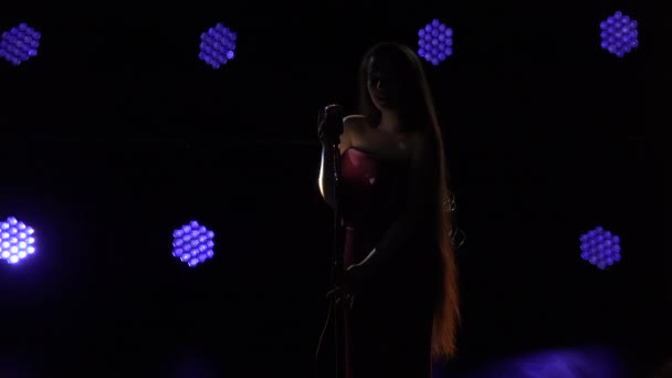 Una bella donna con i capelli lunghi in un abito si erge sul palco cantando in un microfono e si muove sessualmente sullo sfondo di luci blu. Silhouette femminile da vicino. — Video Stock