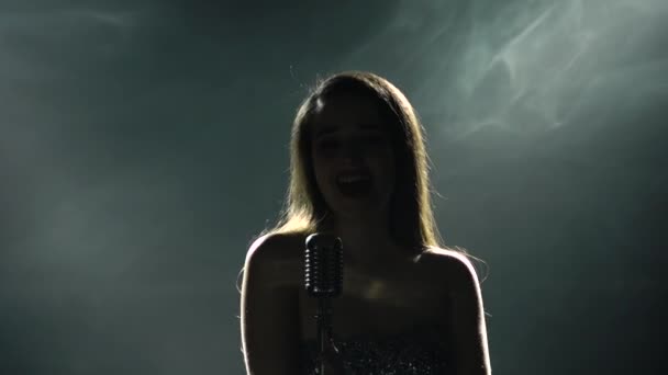 Singing woman artist in a shiny dress on stage. A close-up of the singer in the stage lights and smoke. Young woman singing a song at a live concert into a vintage microphone. Silhouettes. — Stock Video