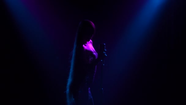 Profesionální zpěvák zpívá ve tmě na jevišti píseň s modrým světlem. Mladá žena v lesklých šatech zpívá do klasického mikrofonu na opravdovém koncertě. Silueta zblízka. — Stock video