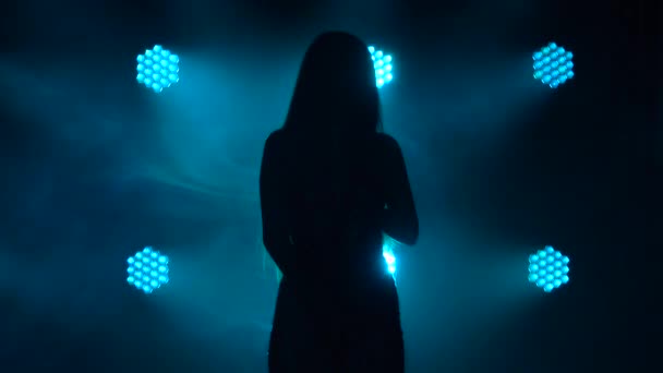 Uma silhueta escura de uma jovem mulher atraente que canta em um microfone vintage. Filmado num estúdio escuro com luz azul nas costas. Fechar. — Vídeo de Stock