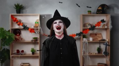 Vampir makyajlı ve cadı şapkalı bir kadın ürkütücü bir Cadılar Bayramı tarzıyla süslenmiş bir odada dikilirken ürkütücü bir şekilde gülüyor. Gotik, fantezi ve tatil teması için mükemmel..