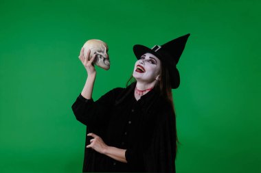 Vampir makyajı ve siyah cadı şapkalı bir kadın yeşil krom anahtar stüdyo arka planında kafatasıyla poz veriyor. Gotik, fantezi ve Cadılar Bayramı teması için mükemmel..