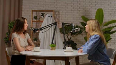 İki kadın mikrofonlu bir masada oturmuş podcast kaydediyorlar. Arka planda hayalet kostümü giymiş bir adam var. Neşeli bir sohbet ve Cadılar Bayramı temalı mizah karışımı..