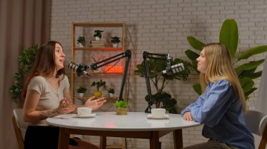 İki kadın, mikrofonlar ve kahve fincanlarıyla yuvarlak bir masada oturur, rahat bir stüdyoda bitki ve neon ışıklı bir dekoru olan bir podcast kaydederler..