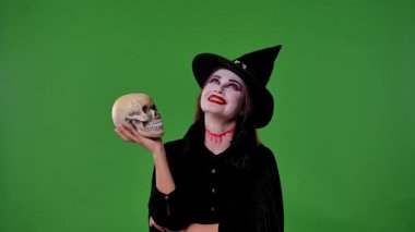 Vampir makyajı ve siyah cadı şapkalı bir kadın yeşil krom anahtar stüdyo arka planında kafatasıyla poz veriyor. Gotik, fantezi ve Cadılar Bayramı teması için mükemmel..