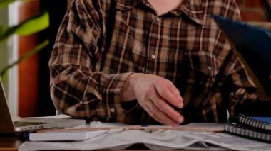 Ekose fanila gömlekli biri dizüstü bilgisayarda çalışıyor, etrafı ahşap bir masada belgelerle çevrili..