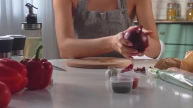Bir kadın, iyi donanımlı bir mutfakta, lezzetli bir yemek hazırlayan bir kesme tahtasında kırmızı soğan dilimliyor. Kırmızı biber, sarımsak, otlar ve baharatlar masaya serilmiştir..