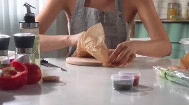 Mutfak ortamında taze sebze ve baharatlarla çevrili bir kesme tahtasındaki tavuğu titizlikle dilimleyen kişi. Evde sağlıklı yemekler hazırlama süreci.