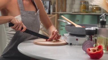 Önlüklü bir kadın mutfaktaki bir kesme tahtasının üzerinde sarımsak dilimliyor. Yanında da yemek malzemeleri var..