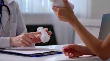 Beyaz önlüklü bir doktor ilaç şişesini muayene ediyor. Masasında tıbbi aletler ve evraklar var..