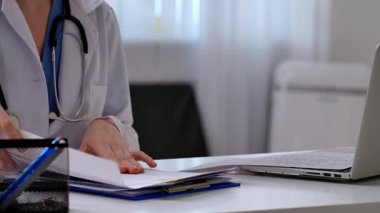 Beyaz önlüklü bir doktor, tıbbi malzemeler ve dizüstü bilgisayarı olan bir reçete yazar..