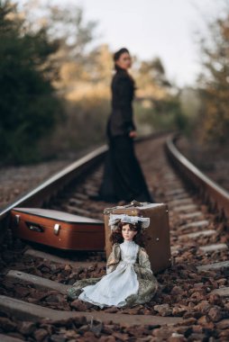 Antika bavullar ve rayların üzerinde eski model oyuncak bebek standı. Arka planda, şık siyah elbiseli bir kadın batan güneşin arka planında duruyor..