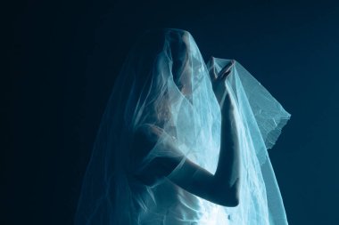 Karanlık bir sahnede tüyler ürpertici bir Cadılar Bayramı sahnesi, şeffaf bir duvakla kaplanmış ölü bir gelin figürü, ürkütücü bir mavi ışıkla aydınlatılmış. Gelinlik ve duvak giymiş bir hayaletin yan görüntüsü..