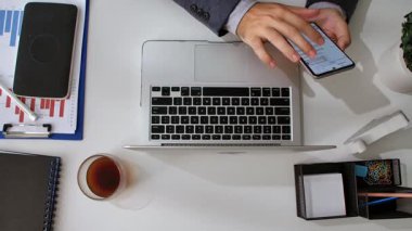 Takım elbiseli bir kişi, ofis malzemeleriyle çevrili bir dizüstü bilgisayarla çalışırken akıllı telefondaki belgeleri inceliyor..