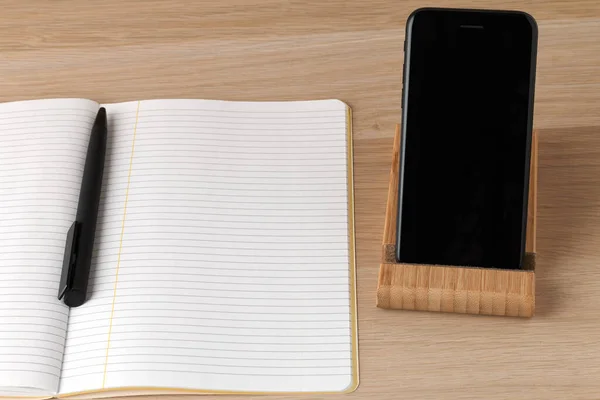 Kurallı bir defter, siyah bir kalem ve siyah bir telefon, çalışma alanı minimalistik.