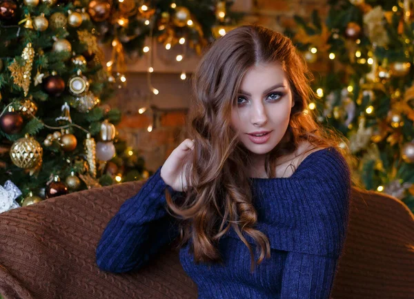Gevşek saçlı güzel bir kız Noel oyuncakları ve çelenklerle bir Noel ağacının yanında oturuyor.