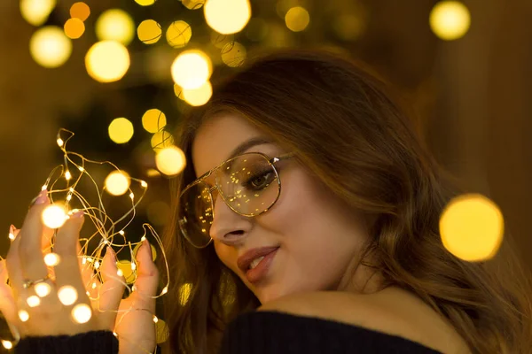 Gevşek saçlı güzel bir kız Noel oyuncakları ve çelenklerle bir Noel ağacının yanında oturuyor.