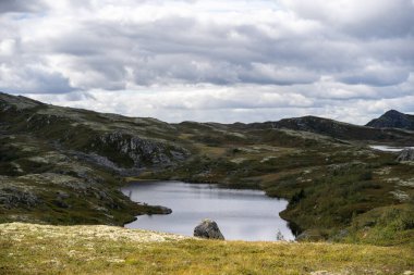 Norveç dağlık kesimlerinde tenha bir dağ gölü manzarası. Sakin, karanlık su dramatik, bulutlu gökyüzünü yansıtır. Huzurlu ve yansıtıcı bir atmosfer yaratır. Etrafı kayalarla çevrili tepelerle çevrili..