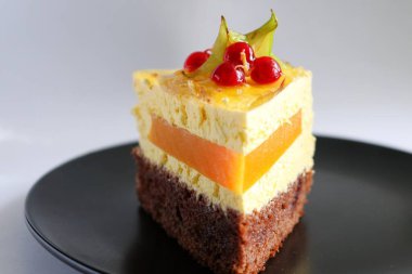Bir dilim mango tutkulu meyveli kek bir dilim karambola ve siyah tabakta kırmızı frenk üzümü ile süslenmiş.