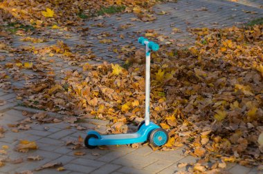 Mavi scooter sonbahar yapraklarının arasında kaldırımın kenarında duruyor.