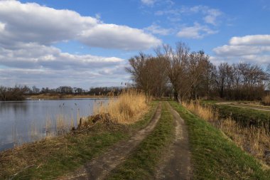 Köprünün geçtiği su kanalı. Etrafta göller ve olgun ağaçlar var. Arka planda bulutlu mavi bir gökyüzü var.