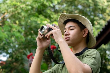 Boonie şapkalı genç bir adam fotoğraf makinesiyle fotoğraf çekiyor. Açık havada çekime odaklanmış ve tutkulu görünüyor..