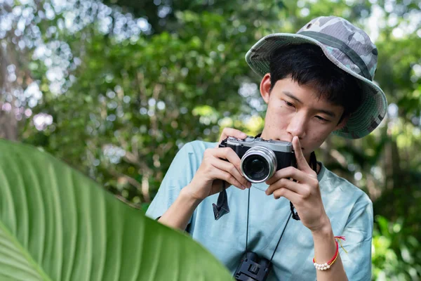 Kamuflaj şapkalı genç bir Asyalı elinde kamerayla sık, yeşil bir ormanda fotoğraf çekiyor. Anı yakalamaya odaklanmış, doğanın ve fotoğrafçılık hobisinin tadını çıkarıyor..