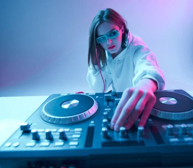 Havalı genç kız DJ karışık konsol ve kulaklıkla müziği karıştırıyor, şık kıyafetler içinde, neon arka planda gözlük takıyor..